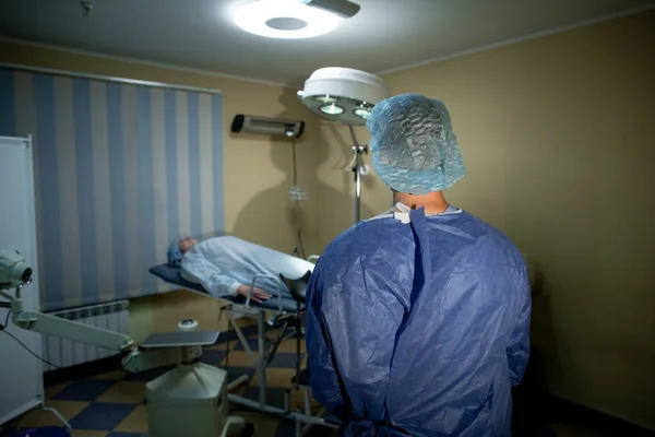 Dokter bedah berdiri dengan punggung ke kamera. Sebelum dia di meja operasi terbaring seorang pasien. Persiapan untuk operasi — Stok Foto