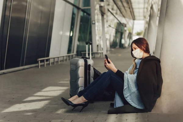 Wanita muda bertopeng medis duduk di dekat bagasi di bandara. Wanita menunggu penerbangan, menggunakan ponsel dan — Stok Foto