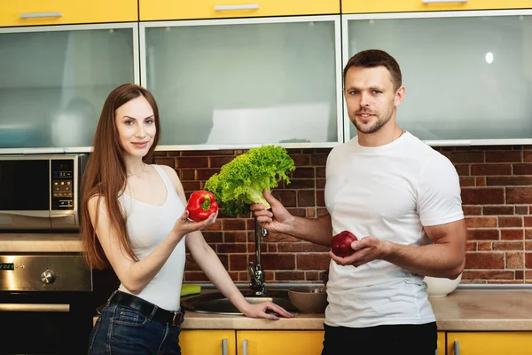 Pasangan muda berdiri di dapur modern dan melihat ke kamera sambil menyiapkan buah-buahan dan sayuran segar. — Stok Foto