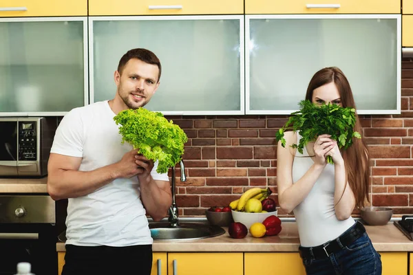 Iklan makanan sehat. Pria dan wanita yang menarik yang berpose di dapur trendi memegang hijau di tangan mereka. Wanita itu menutupi setengah dari wajahnya dengan sekelompok dill dan peterseli — Stok Foto