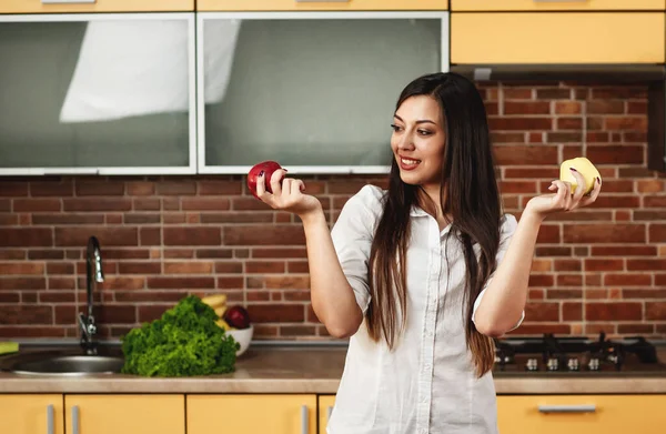 Wanita cantik berambut cokelat berdiri di dapur dan memegang dua apel yang menenangkan di tangannya. — Stok Foto