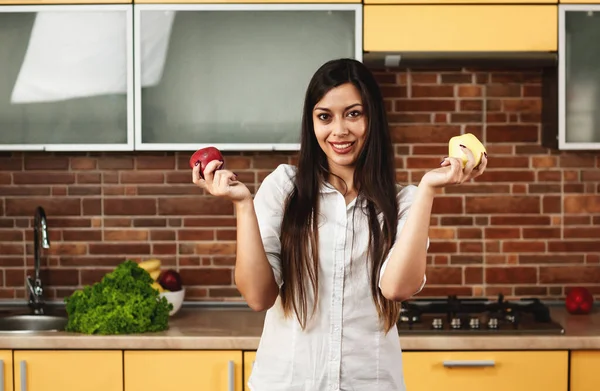 Wanita berambut coklat memegang apel merah dan kuning di tangannya. Seorang wanita berdiri di dapur dan memilih apel mana untuk dimakan. Wanita melihat ke kamera. Konsep diet dan makanan sehat — Stok Foto