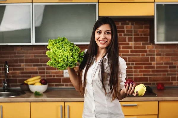 Wanita muda yang bahagia memegang dua apel dan selada di tangannya. Seorang wanita tersenyum dan berdiri di dapur di rumah. — Stok Foto