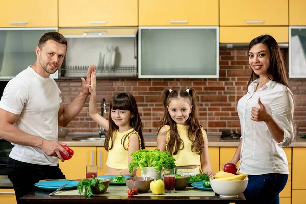Dua gadis kecil yang lucu dan orang tuanya yang cantik memasak sayuran di dapur di rumah. Keluarga bahagia, ayah ibu dan dua anak perempuan, melihat kamera — Stok Foto