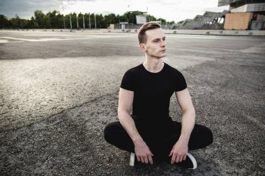 Genç adam asfaltta oturuyor ve koşmadan önce bacaklarını açıyor. Koşmak, spor, sağlıklı yaşam tarzı. Boşluğu kopyala.