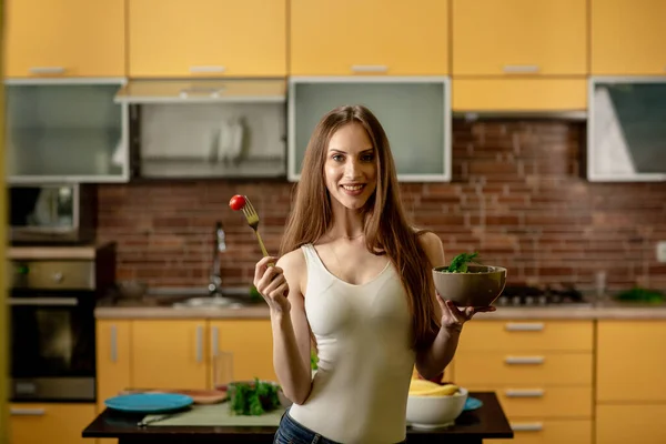 Diet, konsep makan sehat. Wanita muda memegang garpu dengan tomat ceri dan semangkuk salad. Wanita berdiri di dapur di rumah, tersenyum, melihat ke kamera — Stok Foto
