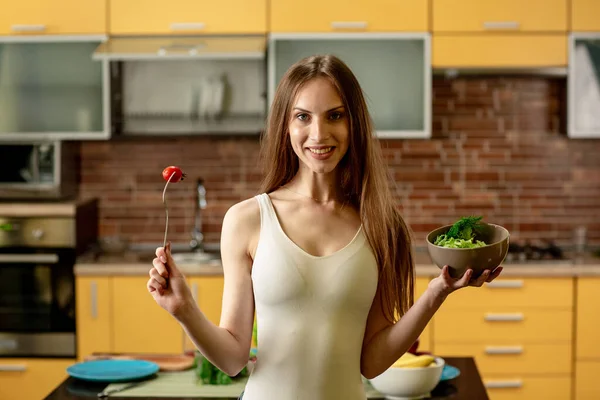 Diet, konsep makan sehat. Wanita muda memegang garpu dengan tomat ceri dan semangkuk salad. Wanita berdiri di dapur di rumah, tersenyum, melihat ke kamera — Stok Foto