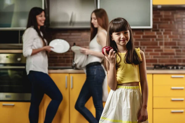 Gadis kecil yang manis memegang apel di tangan. Gadis bahagia berdiri di dapur dan bermain dengan buah-buahan. Di latar belakang, dua wanita menumpuk piring — Stok Foto