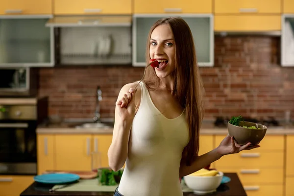 Diet, konsep makan sehat. Wanita muda makan tomat ceri dan memegang semangkuk salad. Wanita berdiri di dapur di rumah, melihat ke kamera — Stok Foto