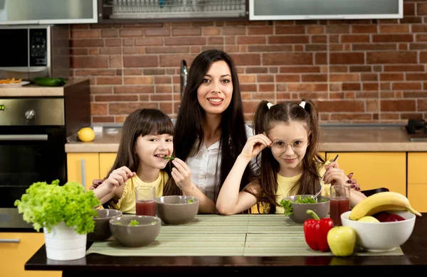Ibu yang ceria dan dua anak perempuan sedang makan salad sehat bersama di rumah. Konsep makan sehat, nilai-nilai keluarga, waktu bersama — Stok Foto