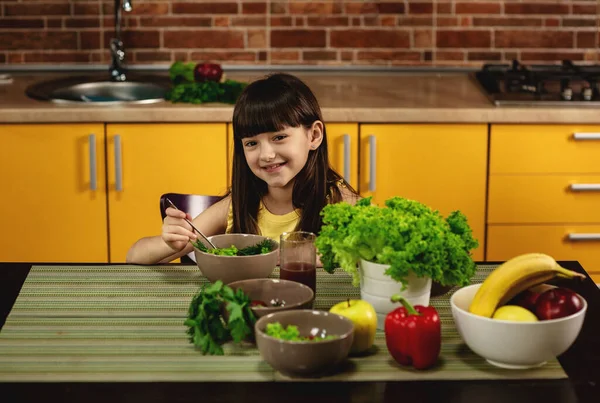 Gadis kecil duduk di meja di dapur dan makan salad. Gadis kecil yang cantik tersenyum. Makanan sehat, sayuran dan buah-buahan — Stok Foto