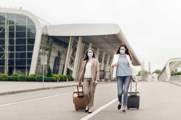 Duas mulheres felizes em máscaras protetoras depois da quarentena do coronavírus com malas vão para o aeroporto. Jovens mulheres perto do aeroporto, abertura de viagens aéreas, conceito de viagem — Fotografia de Stock
