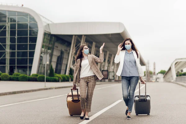 Dua wanita bahagia bertopeng pelindung setelah karantina coronavirus dengan koper pergi ke bandara. Wanita muda di dekat bandara, membuka perjalanan udara, konsep perjalanan — Stok Foto
