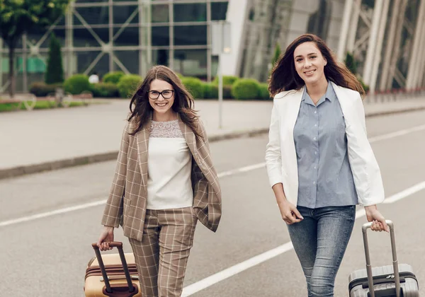 Estilo de vida ao ar livre retratos urbanos de duas mulheres bonitas sorridentes com malas. Melhores amigos whiting para voo com bagagem de mão. Mulheres são enviadas para estudar em outro país — Fotografia de Stock