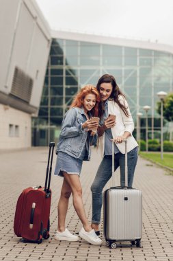 Havaalanında cep telefonuyla arama yapan iki hoş genç kadın. Kadınlar tatile küçük bavullarla geldiler. Seyahat ve rota planlaması