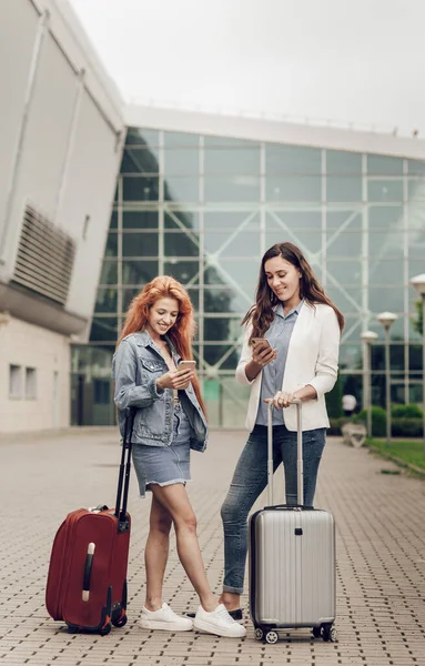 Dua wanita muda yang lucu mencari lokasi di ponsel di bandara. Para wanita datang berlibur dengan koper kecil. Perencanaan perjalanan dan rute — Stok Foto