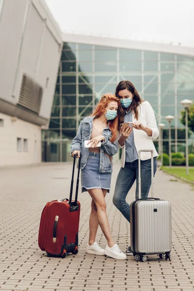 Dua wanita muda bertopeng pelindung berdiri di dekat bandara dan mencari lokasi pada ponsel. Perjalanan setelah karantina coronavirus, pencegahan dan perjalanan yang aman — Stok Foto