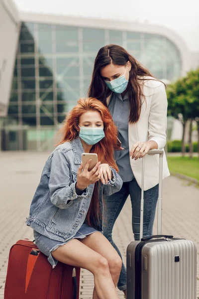 Foto dekat dua wanita muda bertopeng pelindung di dekat bandara. Gadis-gadis mencari lokasi di ponsel. Perjalanan setelah karantina coronavirus, pencegahan dan perjalanan yang aman — Stok Foto