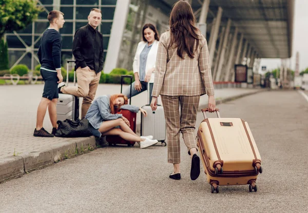 Vista trasera de una mujer con equipaje acercándose a un grupo de amigos. Gente sentada en el asfalto esperando el vuelo. Viajar con amigos, vacaciones —  Fotos de Stock