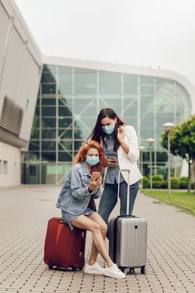 Dua wanita muda bertopeng pelindung berdiri di dekat bandara dan mencari lokasi pada ponsel. Perjalanan setelah karantina coronavirus, pencegahan dan perjalanan yang aman Stok Gambar