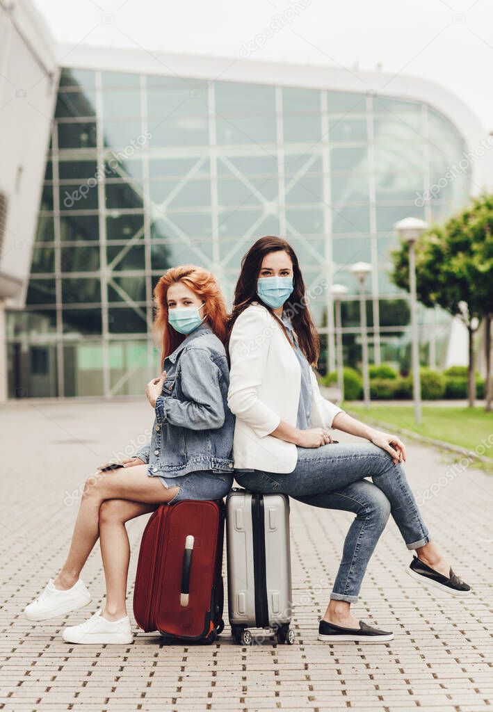 Two women in protective masks sitting on suitcases waiting for the flight. Travel with friends. Tourists waiting for a trip. Travel after quarantine