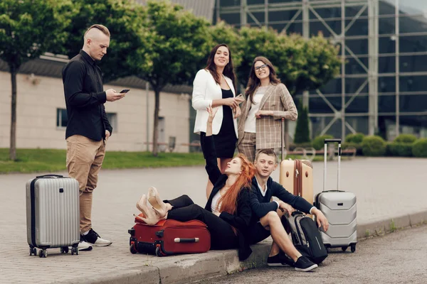 Lima teman menunggu penerbangan, duduk di aspal, menggunakan ponsel. Tiga gadis dan dua anak laki-laki dengan koper pergi pada perjalanan — Stok Foto