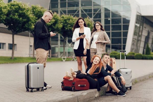 Lima teman menunggu penerbangan, duduk di aspal, menggunakan ponsel. Tiga gadis dan dua anak laki-laki dengan koper pergi pada perjalanan — Stok Foto