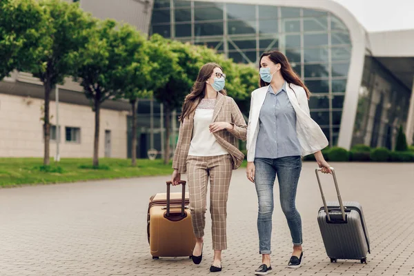보호용 마스크를 쓰고 여행 가방을 든 두 사업가가 공항에 갑니다. 공항 근처의 젊은 여자들이 비행기 여행을 시작하다 로열티 프리 스톡 이미지