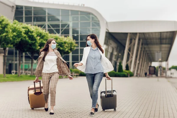 Dua wanita bahagia bertopeng pelindung setelah karantina coronavirus dengan koper pergi ke bandara. Stok Gambar Bebas Royalti
