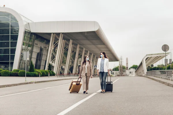 Dua wanita muda dengan koper kembali dari perjalanan bisnis selama karantina. Wanita dengan topeng pelindung Stok Gambar