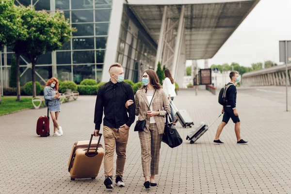 Anak muda dan wanita bertopeng pelindung dengan koper pergi ke bandara. Aman perjalanan setelah pandemi coronavirus. — Stok Foto