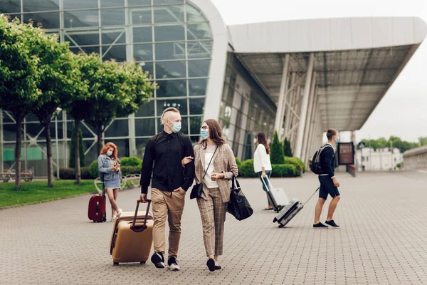 Anak muda dan wanita bertopeng pelindung dengan koper pergi ke bandara. Aman perjalanan setelah pandemi coronavirus. — Stok Foto