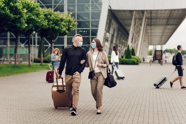 Anak muda dan wanita bertopeng pelindung dengan koper pergi ke bandara. Aman perjalanan setelah pandemi coronavirus. — Stok Foto