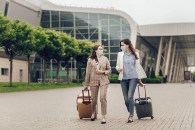 Koruyucu maskeli ve bavullu iki iş kadını havaalanına gidiyor. Havaalanı yakınında genç kadınlar, hava yolculuğu, seyahat konsepti. Koronavirüs karantinası sırasında iş seyahatleri