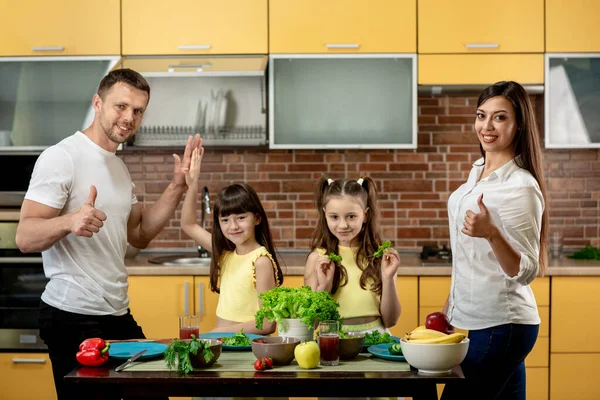 Das Konzept der gesunden Ernährung. Eine junge glückliche Familie mit zwei Töchtern steht am Tisch und bereitet ein gesundes Abendessen zu. Eltern zeigen Handzeichen super — Stockfoto
