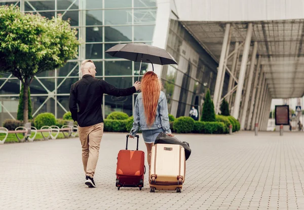 Pasangan bahagia bepergian. Pandangan belakang, wanita memegang dua koper, pria memegang payung, hari hujan — Stok Foto