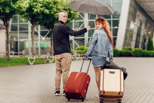 แฮปปี้ คู่รักเดินทาง มุมมองด้านหลัง ผู้หญิงถือกระเป๋าเดินทางสองใบ ผู้ชายถือร่ม วันที่ฝนตก — ภาพถ่ายสต็อก