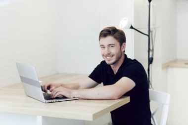 Evde dizüstü bilgisayarla çalışan gülümseyen beyaz bir adam masada oturuyor. Koronavirüs karantinası sırasında belli bir mesafeden çalış. Evde kal, kendini izole et.