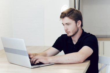 Evde çalışan, dizüstü bilgisayarda yazı yazan genç bir adam masasında oturuyor. Koronavirüs karantinası sırasında belli bir mesafeden çalış. Evde kal, kendini izole et.