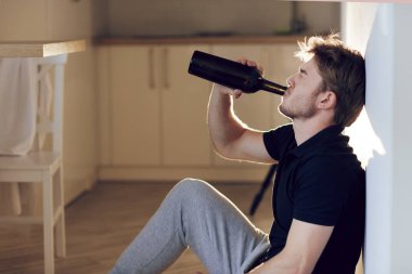Yan manzara, elinde bir şişe şarapla mutfakta yerde oturan sarhoş genç adam. Yalnız içmek. Erkek alkolizmi, kötü alışkanlıklar, depresyon