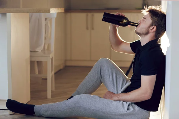 Pandangan samping, pemuda mabuk dengan botol anggur duduk di lantai dapur. Minum sendirian saja. Pria konsep alkoholisme, kebiasaan buruk, depresi — Stok Foto