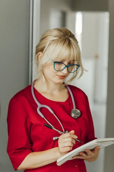Potret close-up seorang dokter muda berambut pirang dengan seragam merah dengan stetoskop. Seorang dokter berkacamata ada di rumah sakit dan menulis sesuatu di buku catatan — Stok Foto