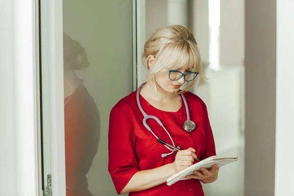 Dokter pirang muda berseragam merah dengan stetoskop. Seorang dokter berkacamata ada di rumah sakit dan menulis sesuatu di buku catatan — Stok Foto