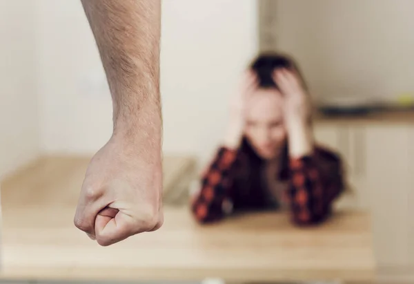 Puño masculino de cerca en foco. Sobre un fondo borroso, una mujer está sentada en una mesa, apretando la cabeza con las manos. Víctima de violencia doméstica — Foto de Stock