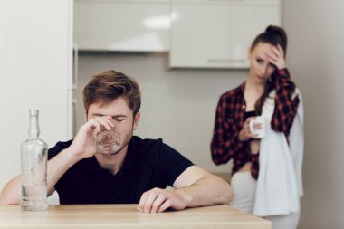 Mutfakta masada oturan bir adam, üzgün bir kadın, bir erkeğin yanında durup ona bakar.
