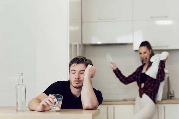 Minum masalah orang mabuk dalam keluarga muda. Seorang pria minum sambil duduk di meja di dapur, seorang wanita di belakangnya — Stok Foto