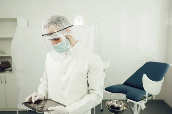 Seorang ginekolog laki-laki dengan tablet di tangan, dalam setelan pelindung mempersiapkan diri untuk menerima pasien selama karantina koronavirus. Dokter dalam peralatan perlindungan pribadi selama pandemi — Stok Foto