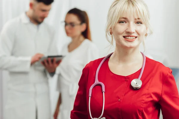 Potret close-up seorang dokter wanita tersenyum dalam seragam merah dengan stetoskop. Dokter muda tersenyum dalam berpose di kamera, di belakangnya di latar belakang dua dokter lainnya — Stok Foto