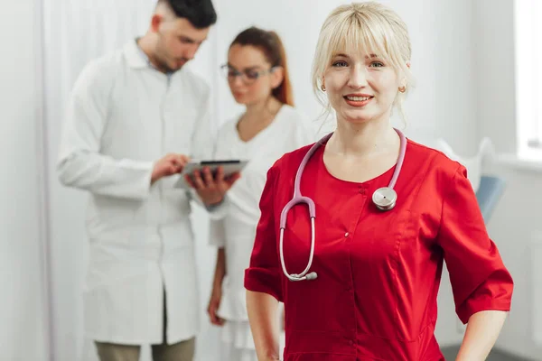 Potret close-up seorang dokter wanita tersenyum dalam seragam merah dengan stetoskop. Dokter muda tersenyum dalam berpose di kamera, di belakangnya di latar belakang dua dokter lainnya — Stok Foto