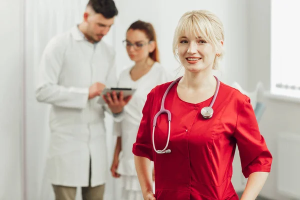 Potret close-up seorang dokter wanita tersenyum dalam seragam merah dengan stetoskop. Dokter muda tersenyum dalam berpose di kamera, di belakangnya di latar belakang dua dokter lainnya — Stok Foto